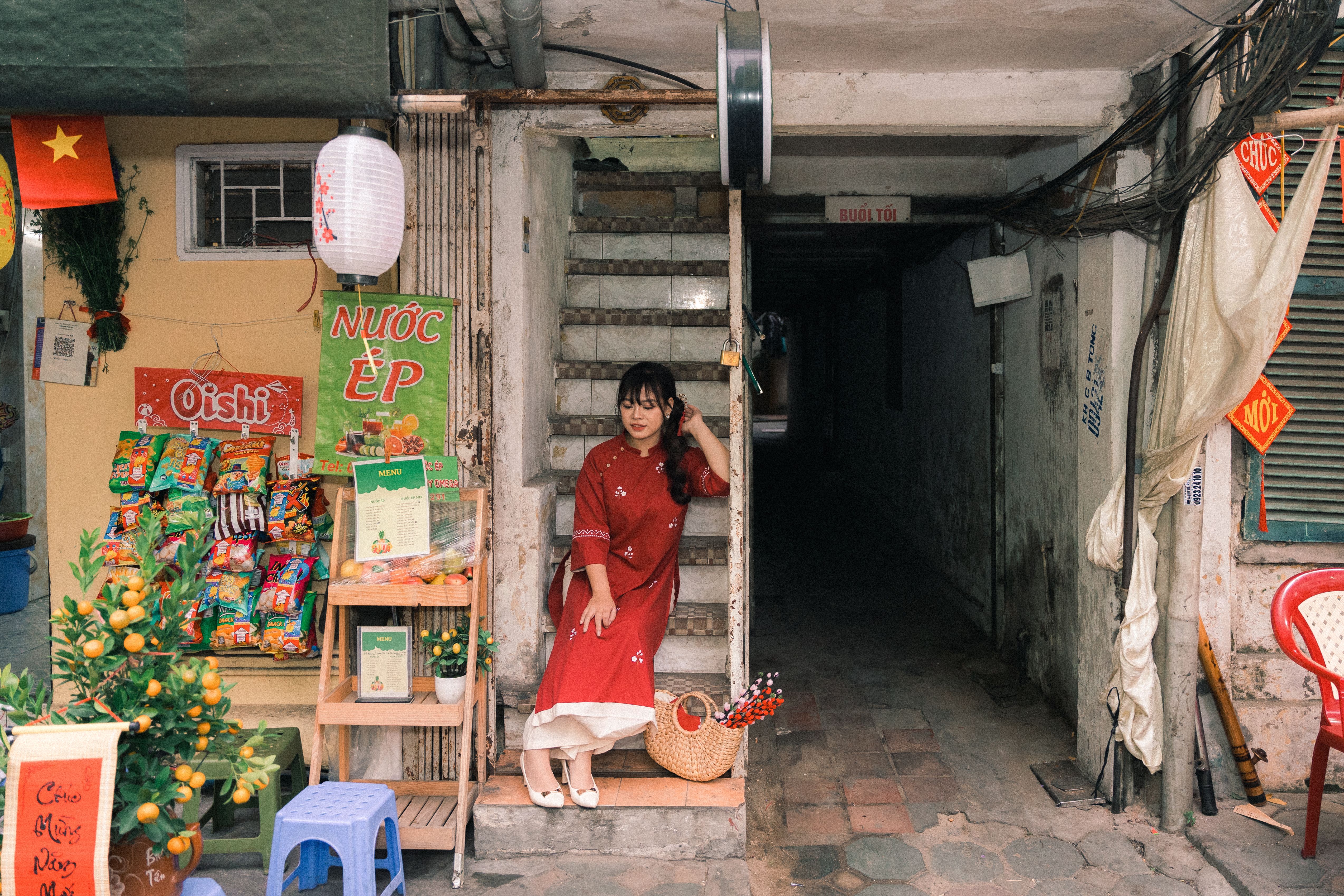 A woman in red dress sits by a shop in Hanoi, Vietnam, surrounded by snacks and decorations.