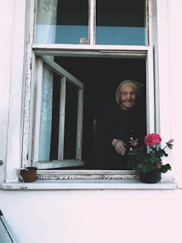 A senior woman stands at an open window with pink flowers, radiating nostalgia.