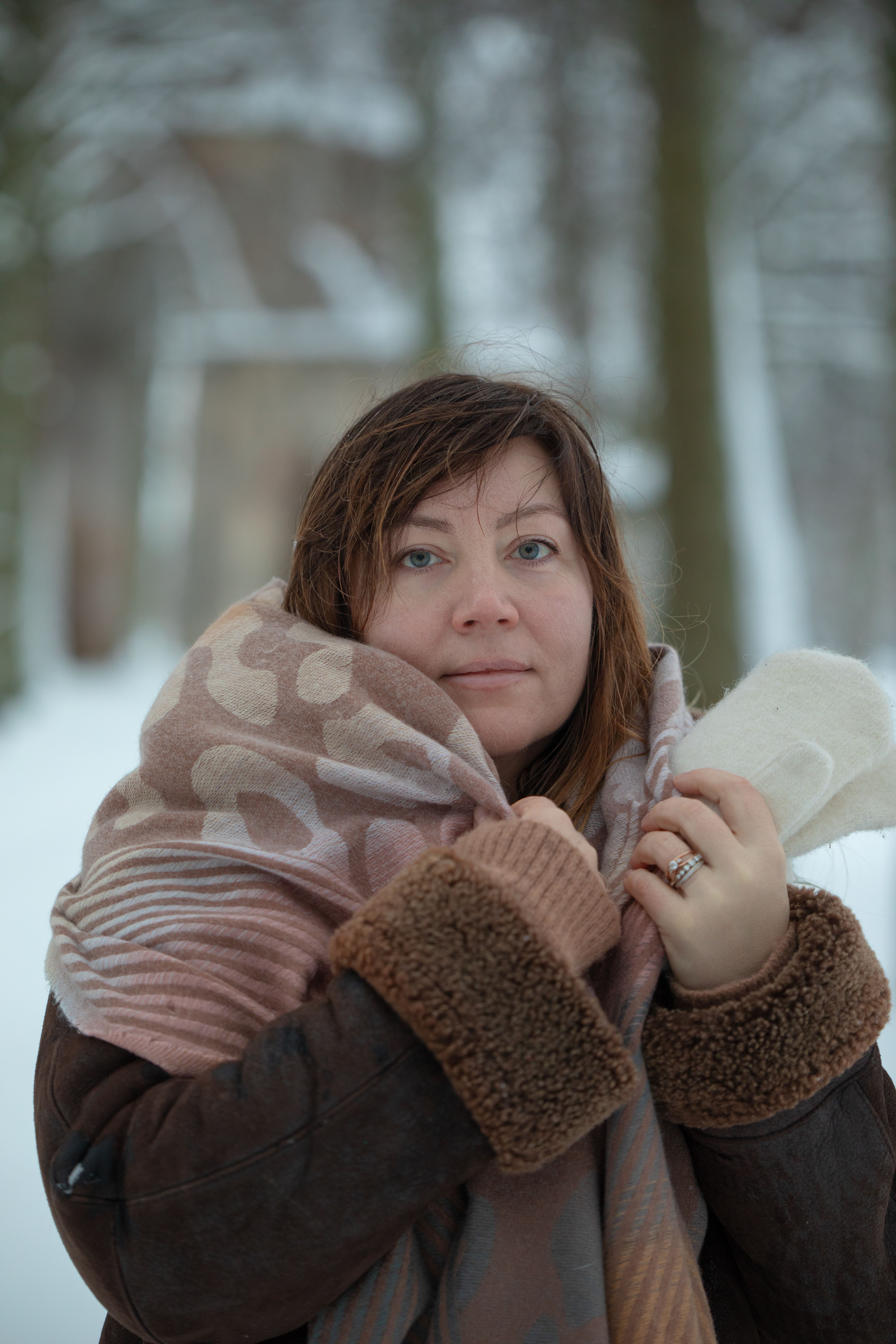 A woman in warm winter clothing stands outdoors in a snowy forest setting, looking towards the camera.