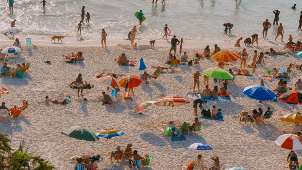 A crowded beach scene with colorful umbrellas and people relaxing in the sunlight.
