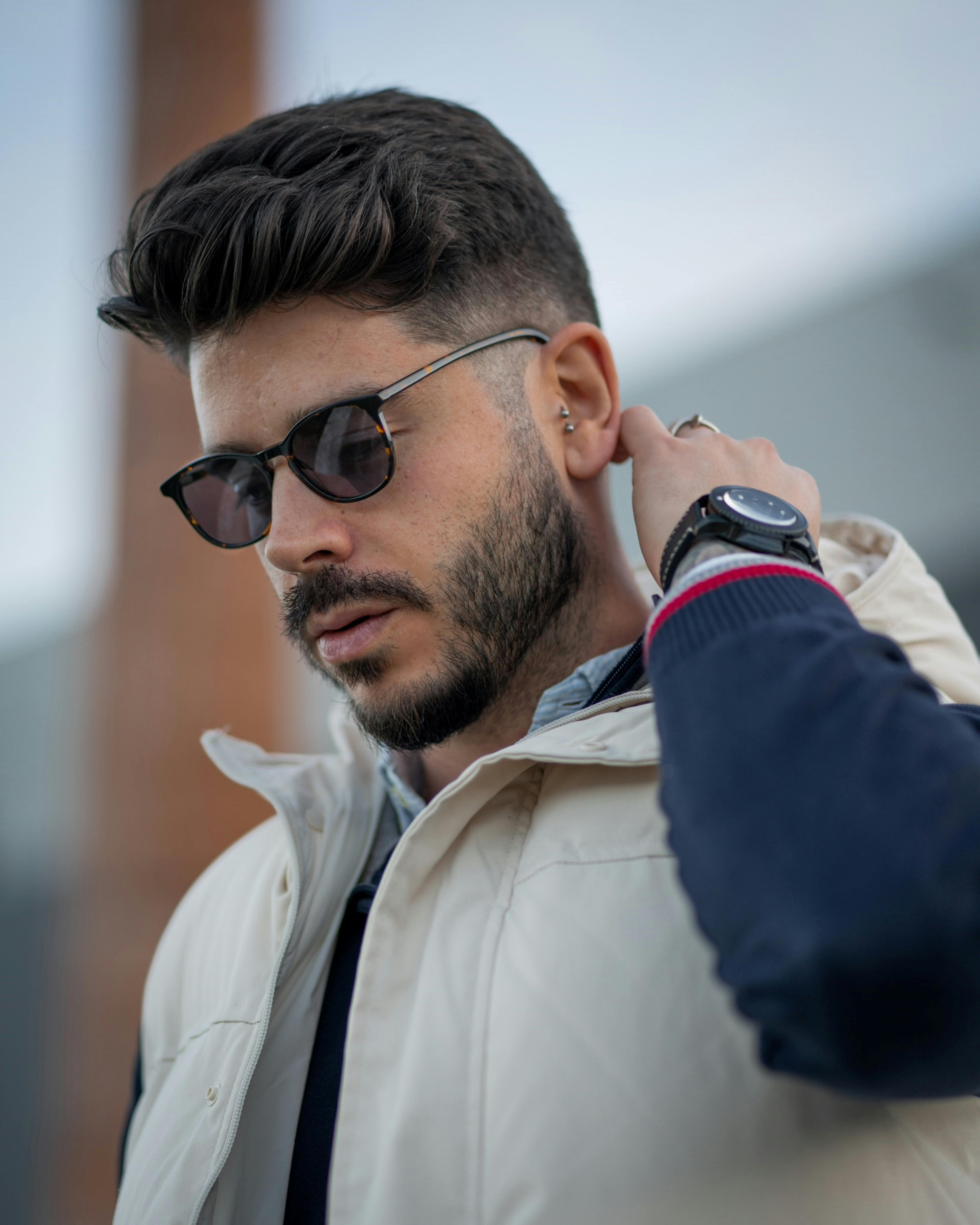 Fashionable male model wearing sunglasses and a watch, posing outdoors with an urban backdrop.