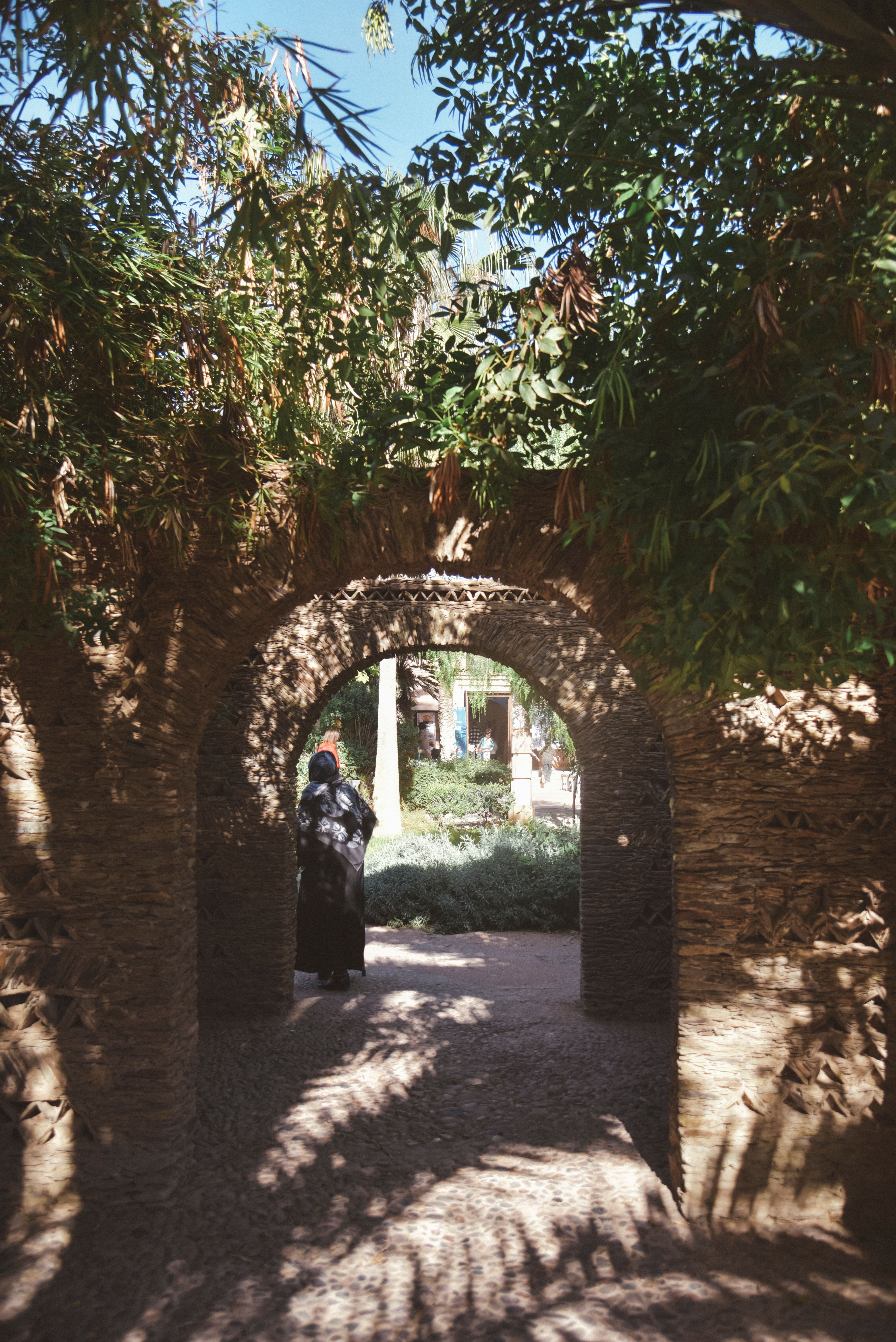 Woman Walking Through Arched Stone Path in Agadir Garden · Free Stock Photo