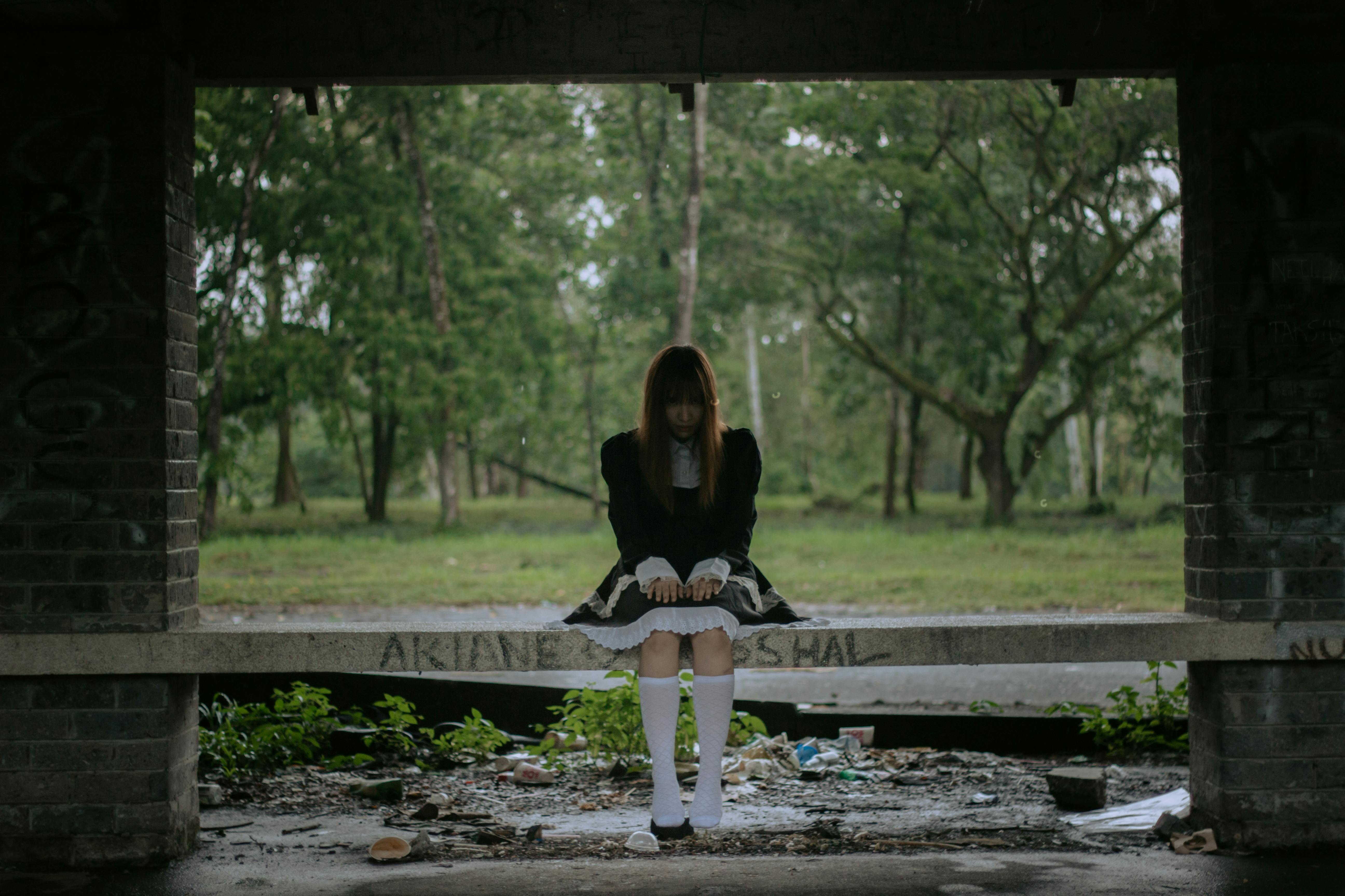 一位身穿黑色连衣裙的年轻女子坐在森林里的废弃长凳上，营造出喜怒无常的氛围