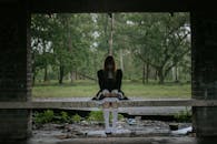 一位身穿黑色连衣裙的年轻女子坐在森林里的废弃长凳上，营造出喜怒无常的氛围