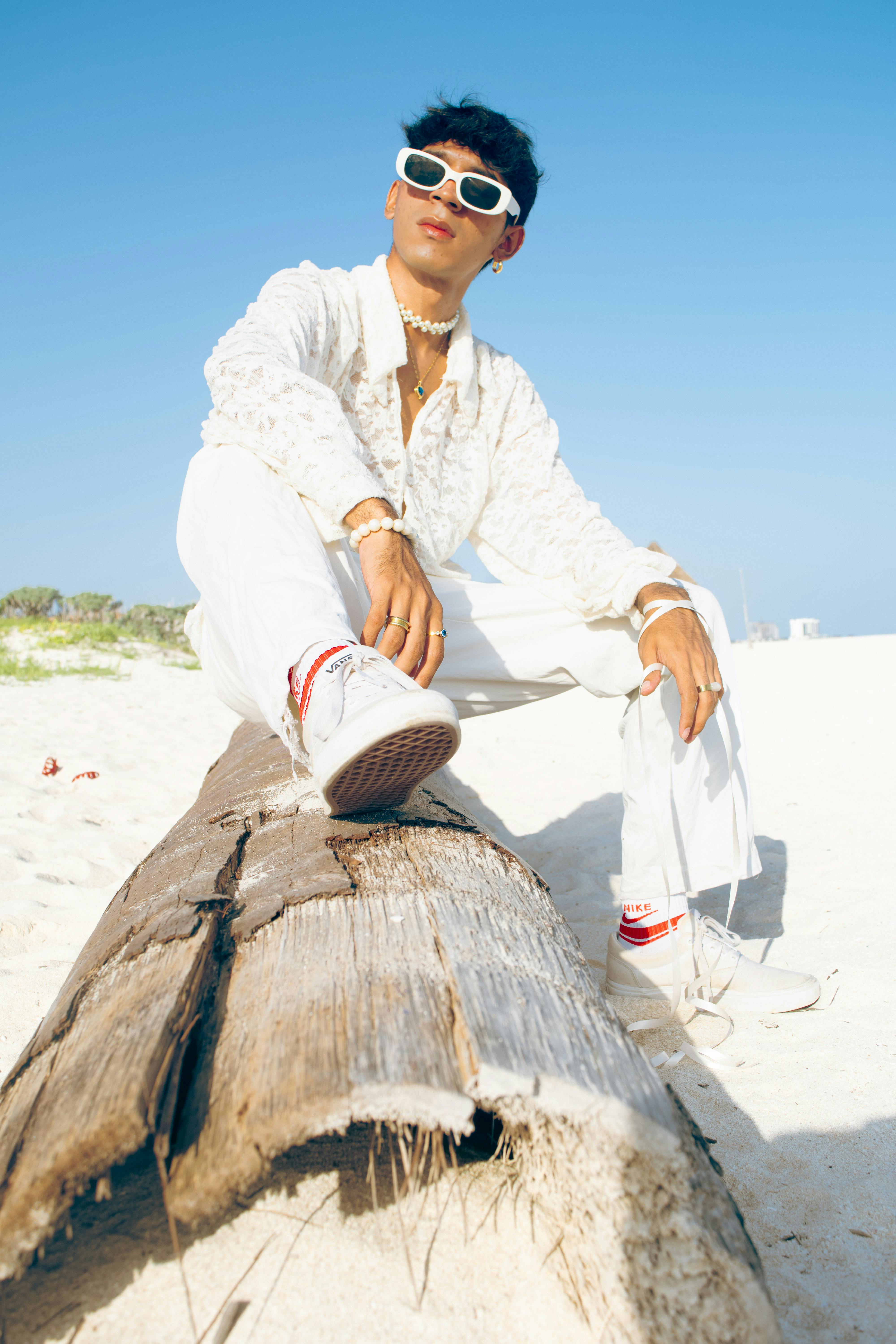 A fashionable adult wearing sunglasses and white attire, posing on beach driftwood under a clear sky.