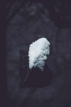 A dark and moody image of a single rock adorned with snow, floating in calm waters.