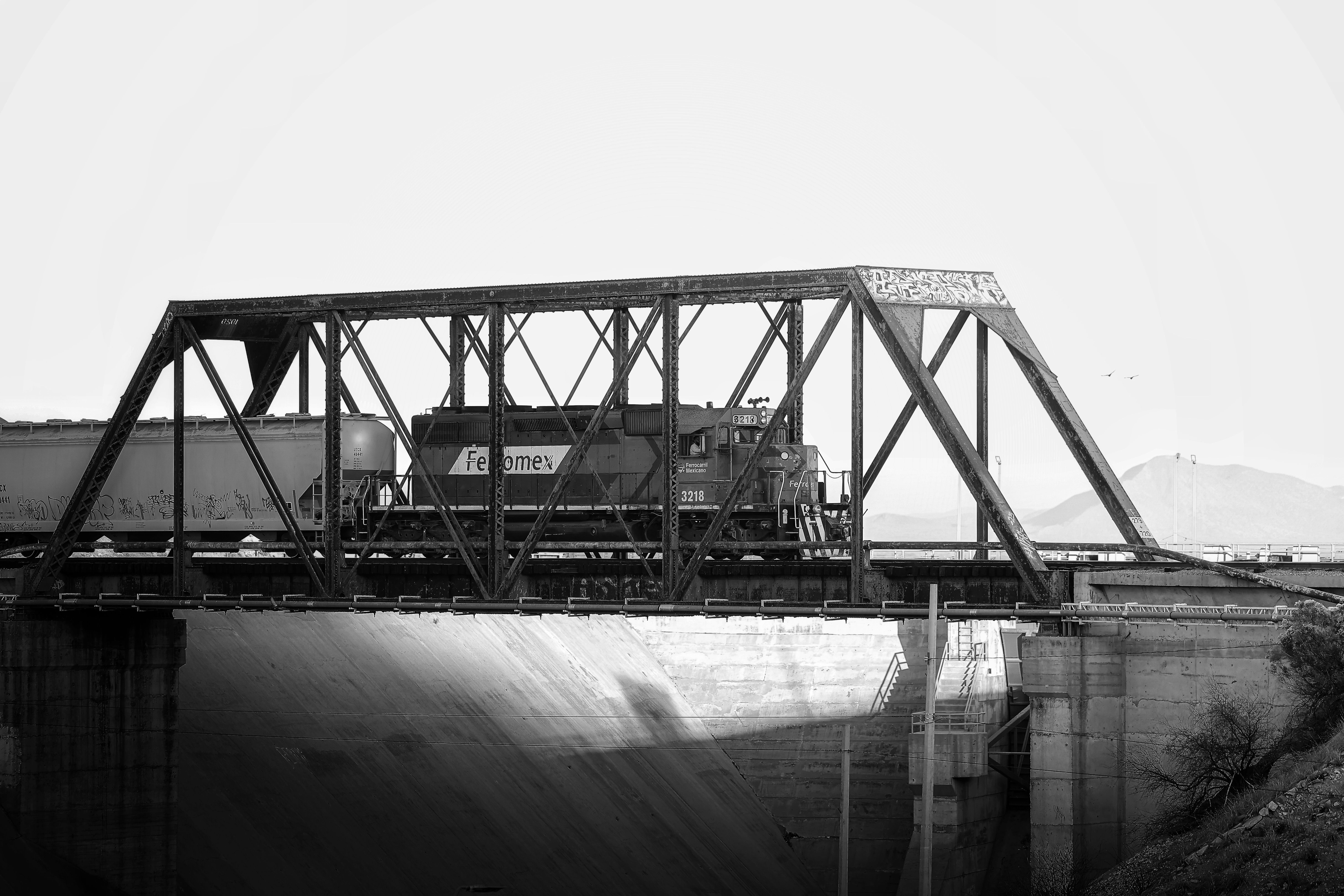 A black and white image of a vintage train crossing a steel bridge with mountains in the background.
