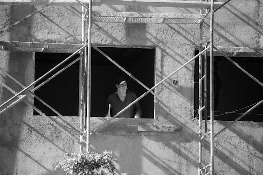A construction worker stands on a building site scaffolding in a black and white photo.