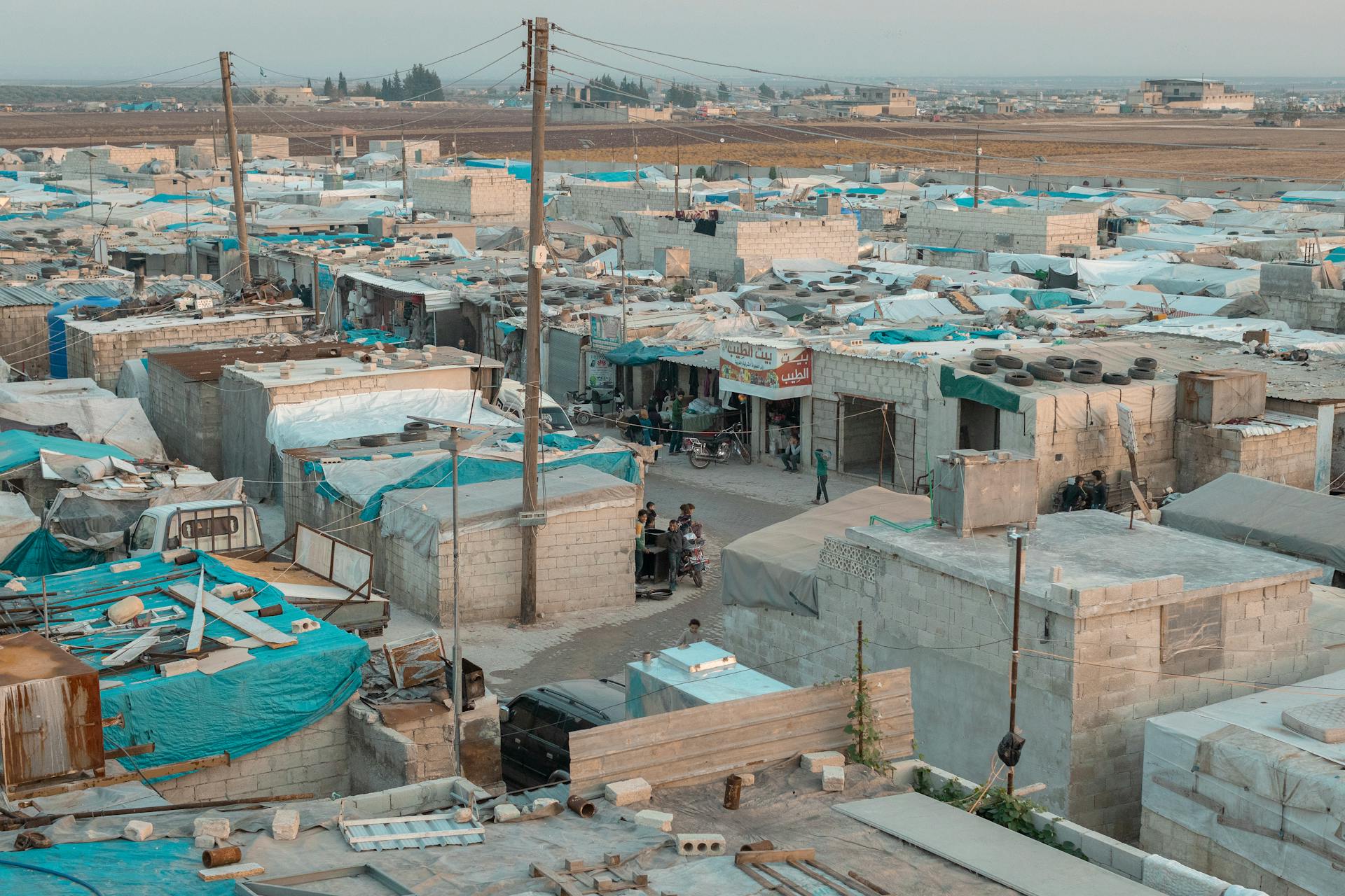 A broad aerial view of a refugee camp with makeshift shelters and blue tarps in an urban area.