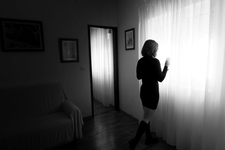 A woman stands by a window with curtains, in a moody black and white room.