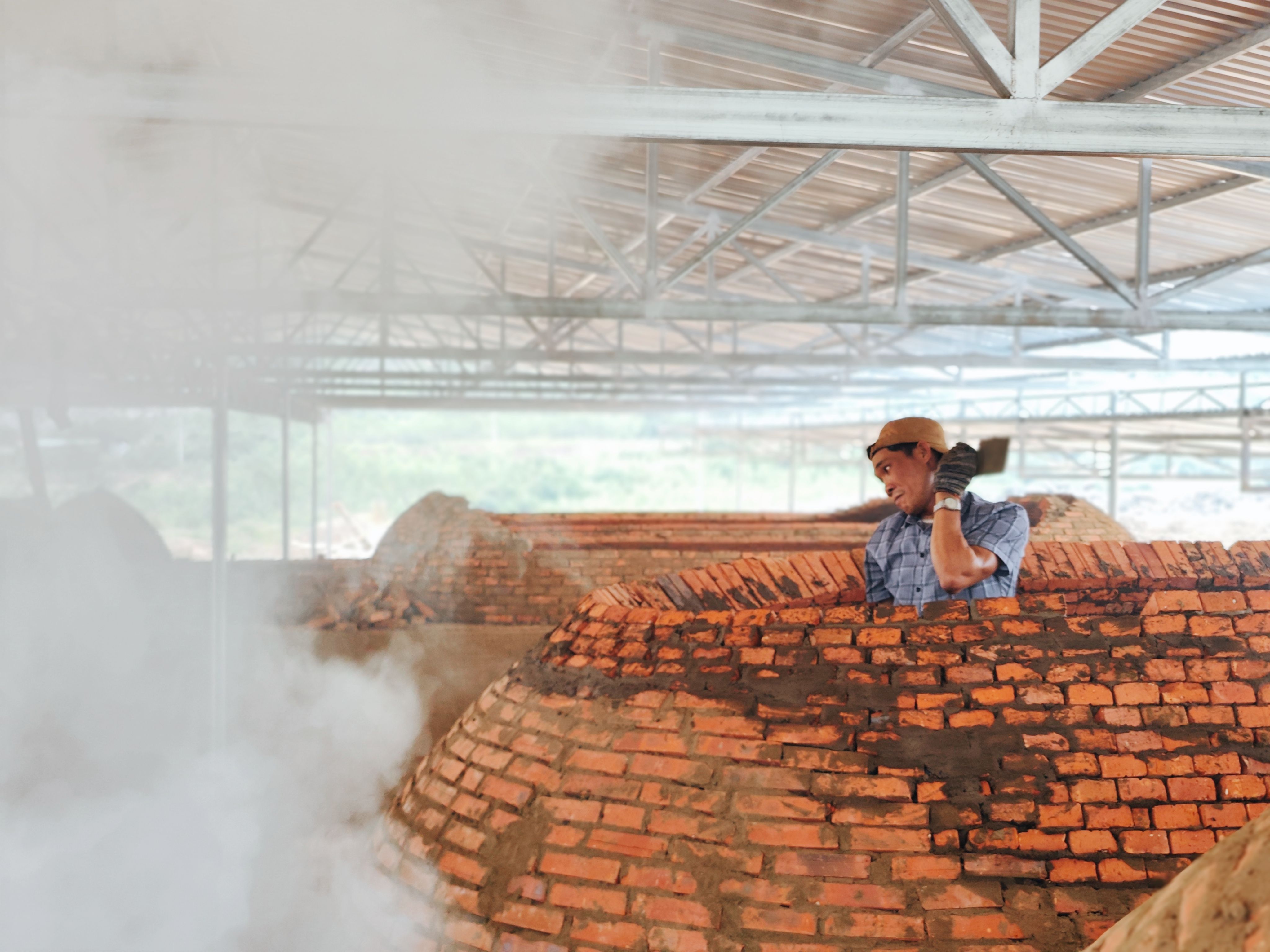 A laborer working among brick kilns with steam rising in Phú Yên, Vietnam, capturing industrial life.