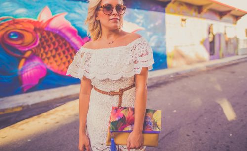 Free Fashionable woman in a lace dress with a colorful clutch stands near a lively street mural. Stock Photo