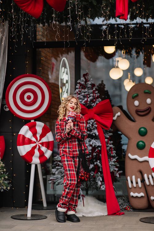 Free Woman in plaid pajamas enjoying holiday decorations outside a cafe. Christmas spirit with gingerbread and candy props. Stock Photo