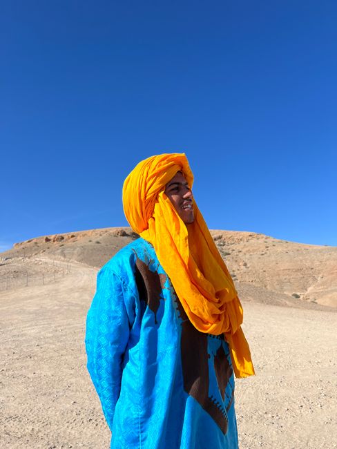 A man in vibrant traditional attire stands in a Moroccan desert landscape under a clear blue sky.