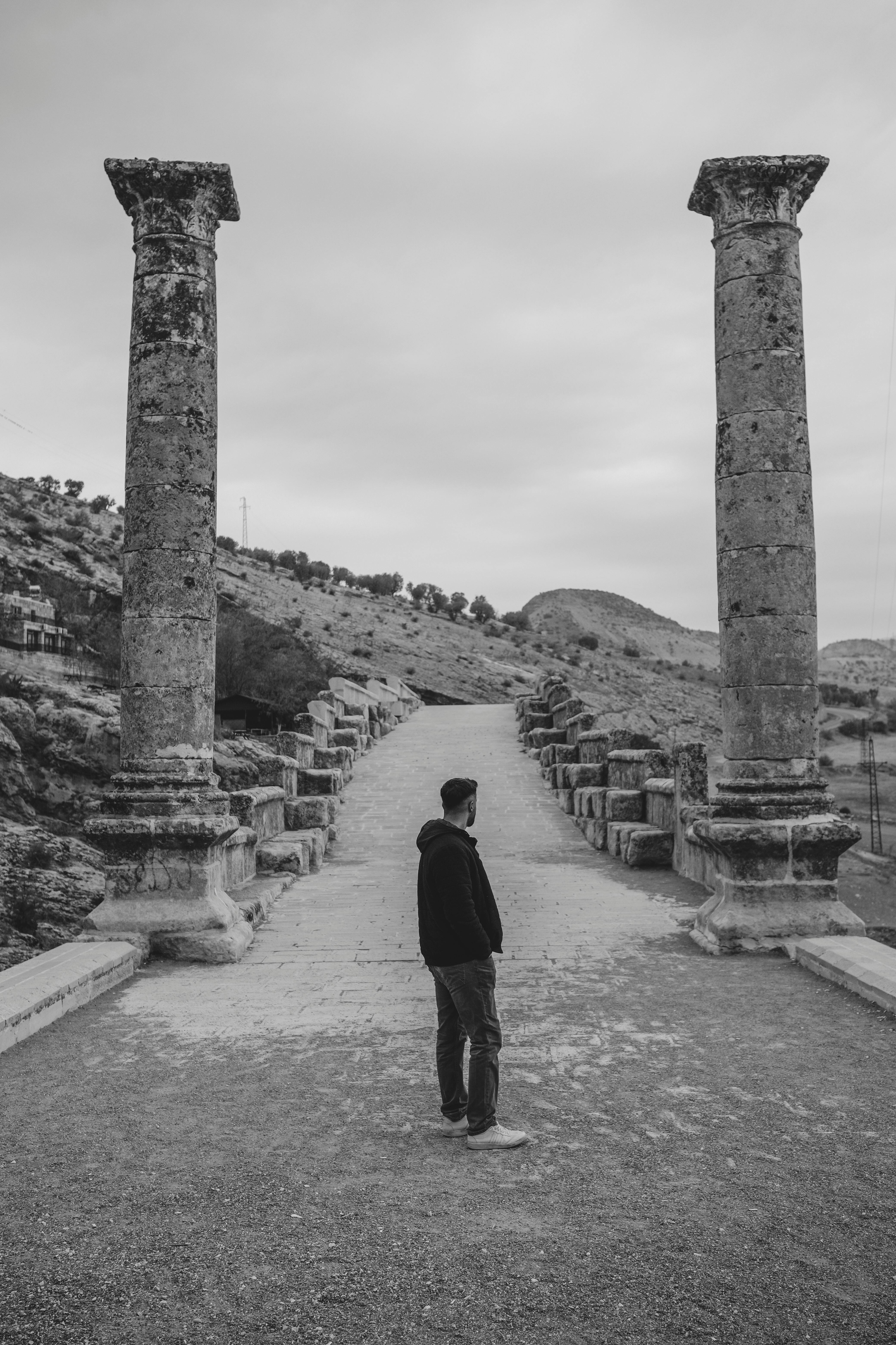 A lone person stands between ancient pillars on a historic path in black and white.