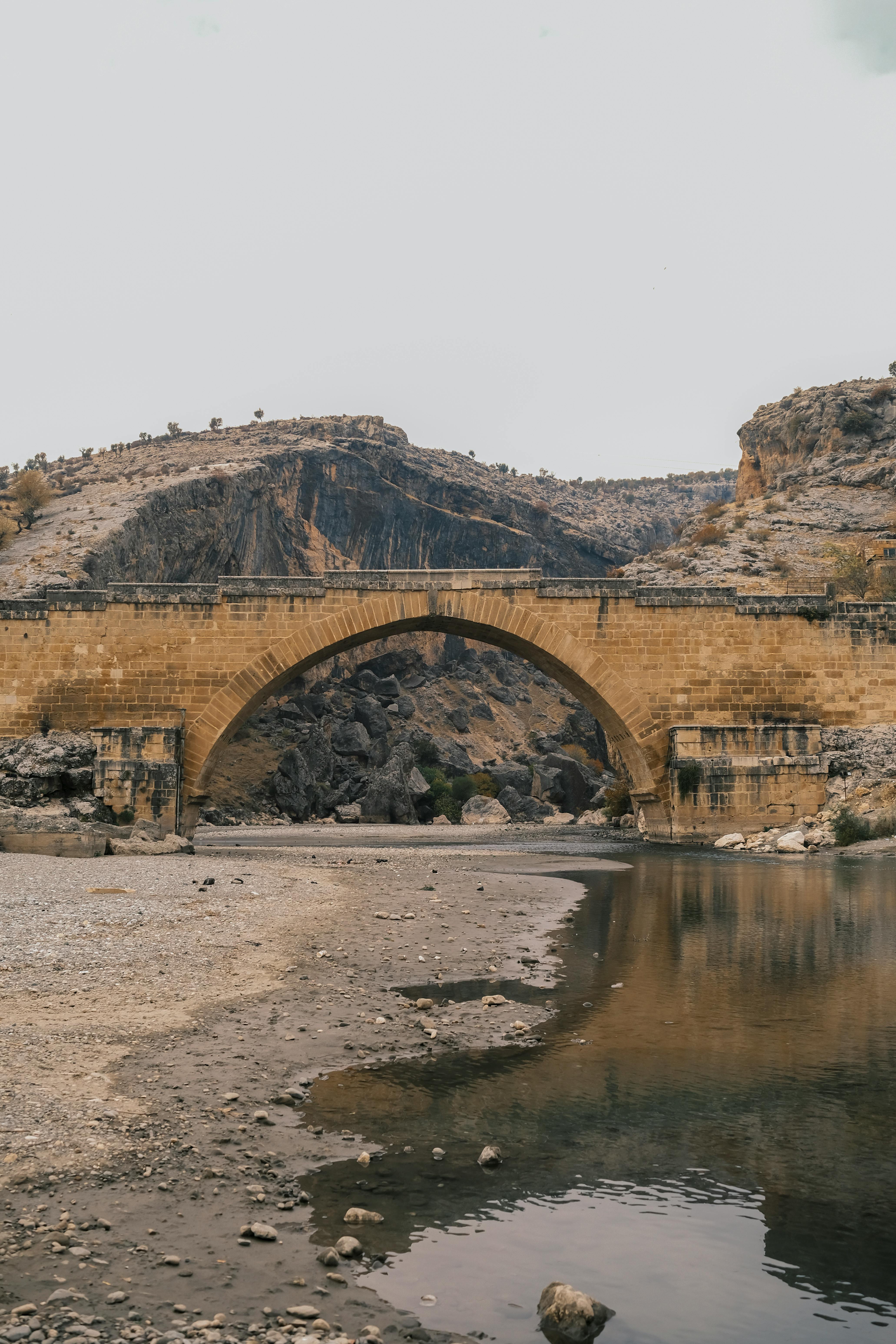 Explore the timeless beauty of an ancient stone bridge over a tranquil river in a rugged landscape.