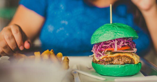 Colorful burger with green bun and fries next to a person's hand, creating a vivid dining scene.