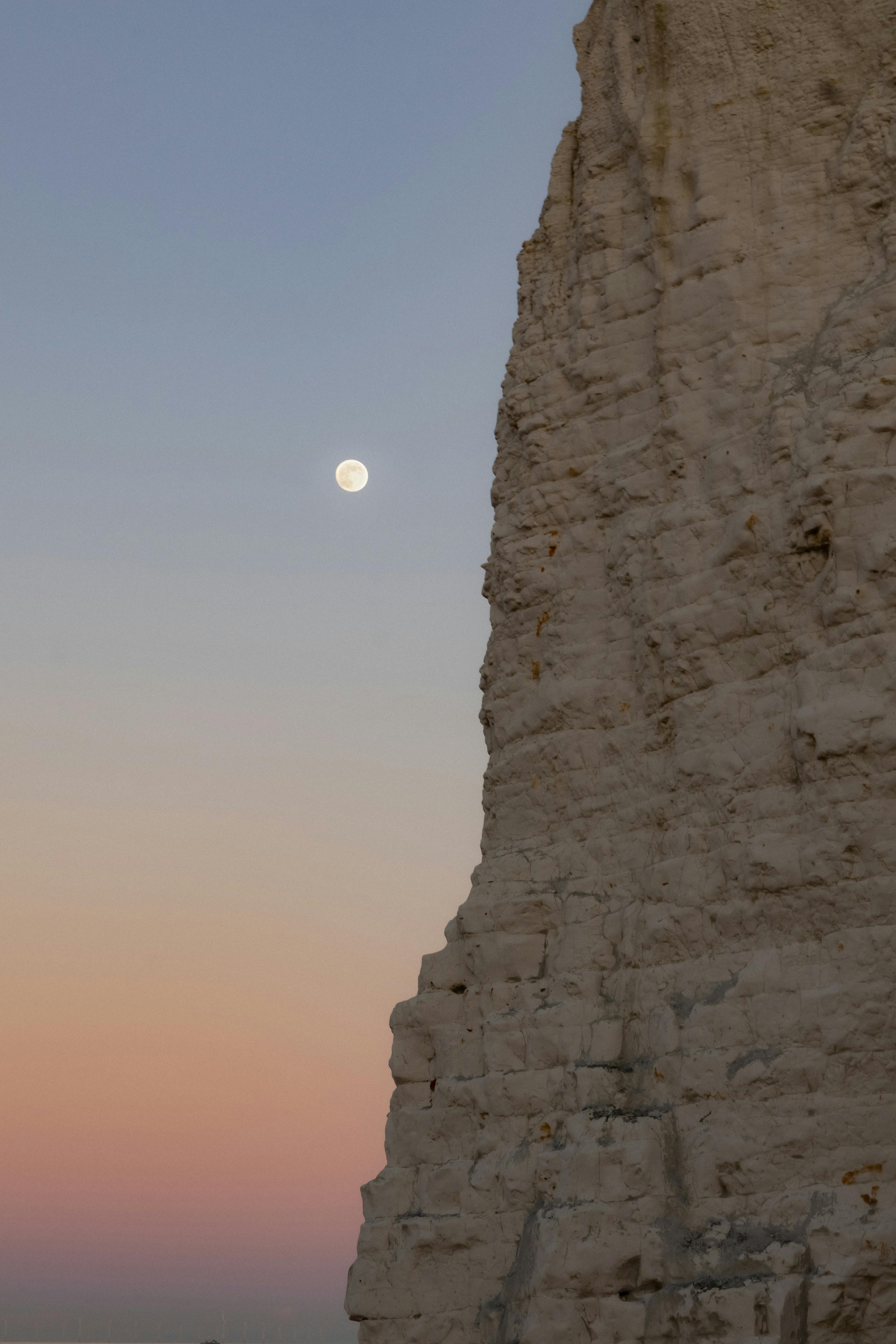 A full moon rises over a rugged cliff at sunset by the sea in a serene landscape.