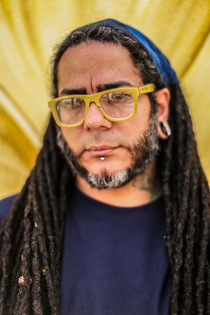 Close-up portrait of a man wearing eyeglasses and dreadlocks, standing against a yellow background.