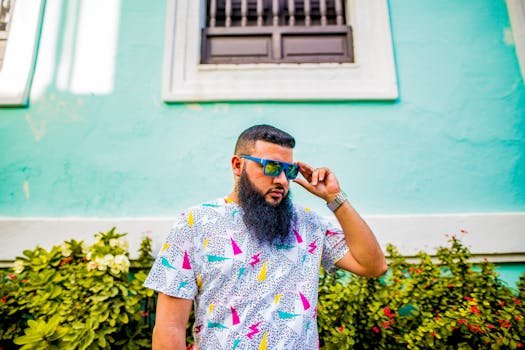 Stylish bearded man wearing sunglasses in front of a vibrant teal wall on a sunny day.