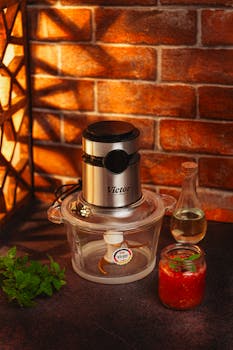 Compact metal blender with glass jar, sauce bottle, and mint on a dark counter.