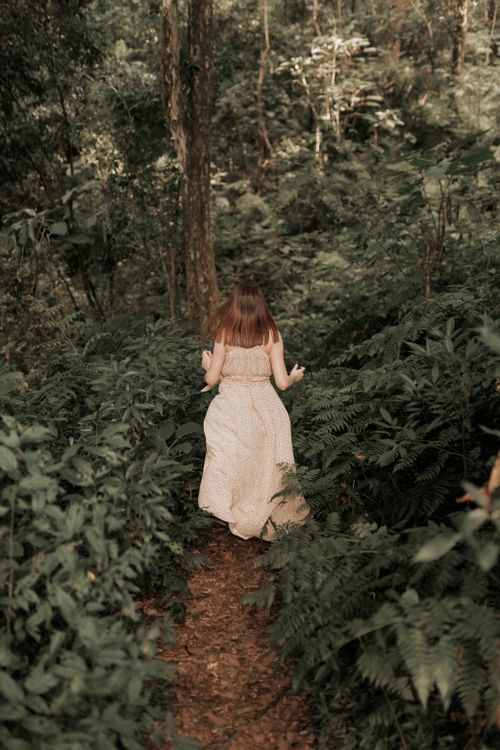 Free A young girl in a white dress explores a dense forest path, surrounded by lush greenery. Stock Photo