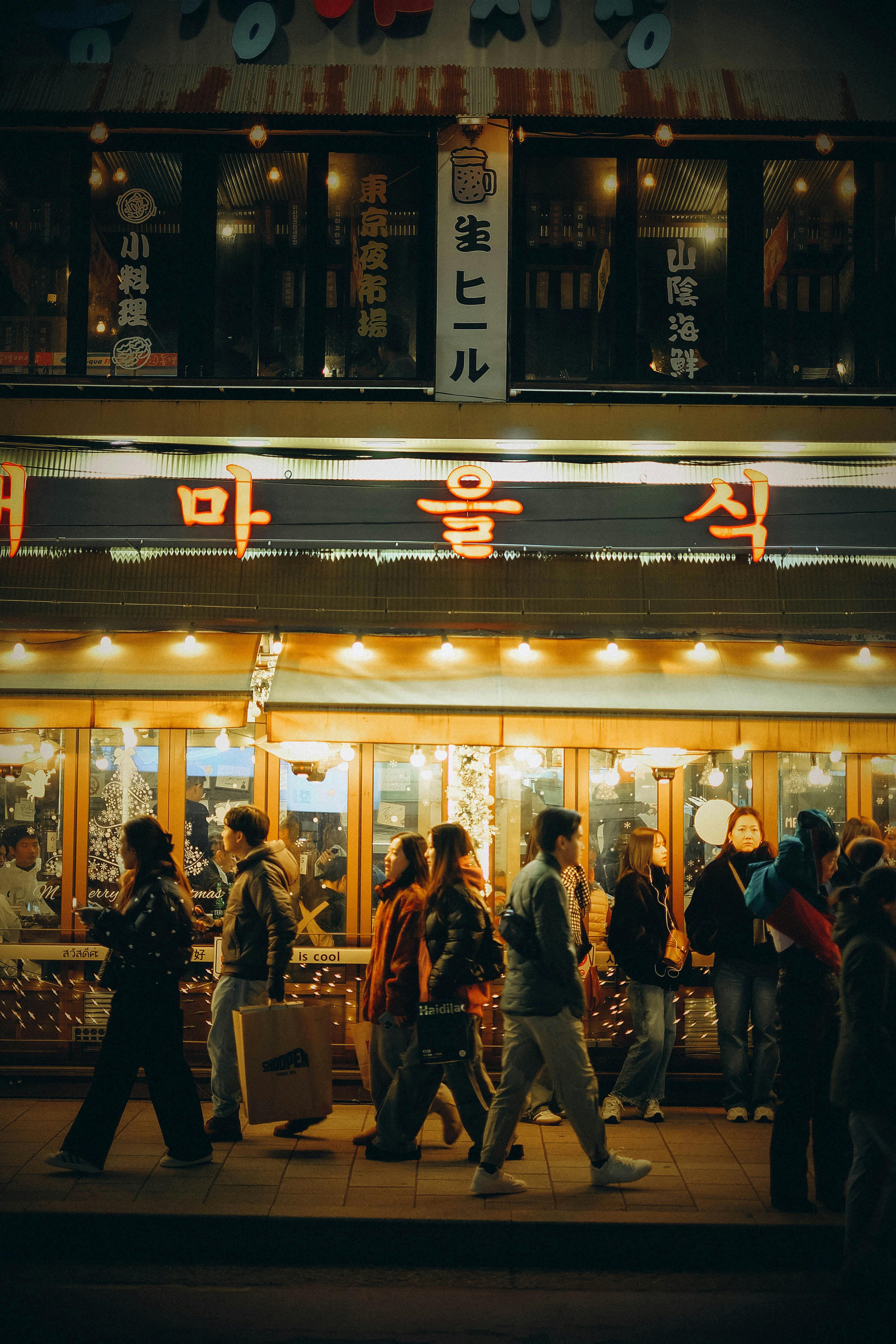 People walking past a lit-up storefront on a busy street in Seoul at night.