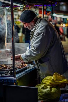 Elderly man roasting chestnuts at a vibrant night market, surrounded by urban lights.