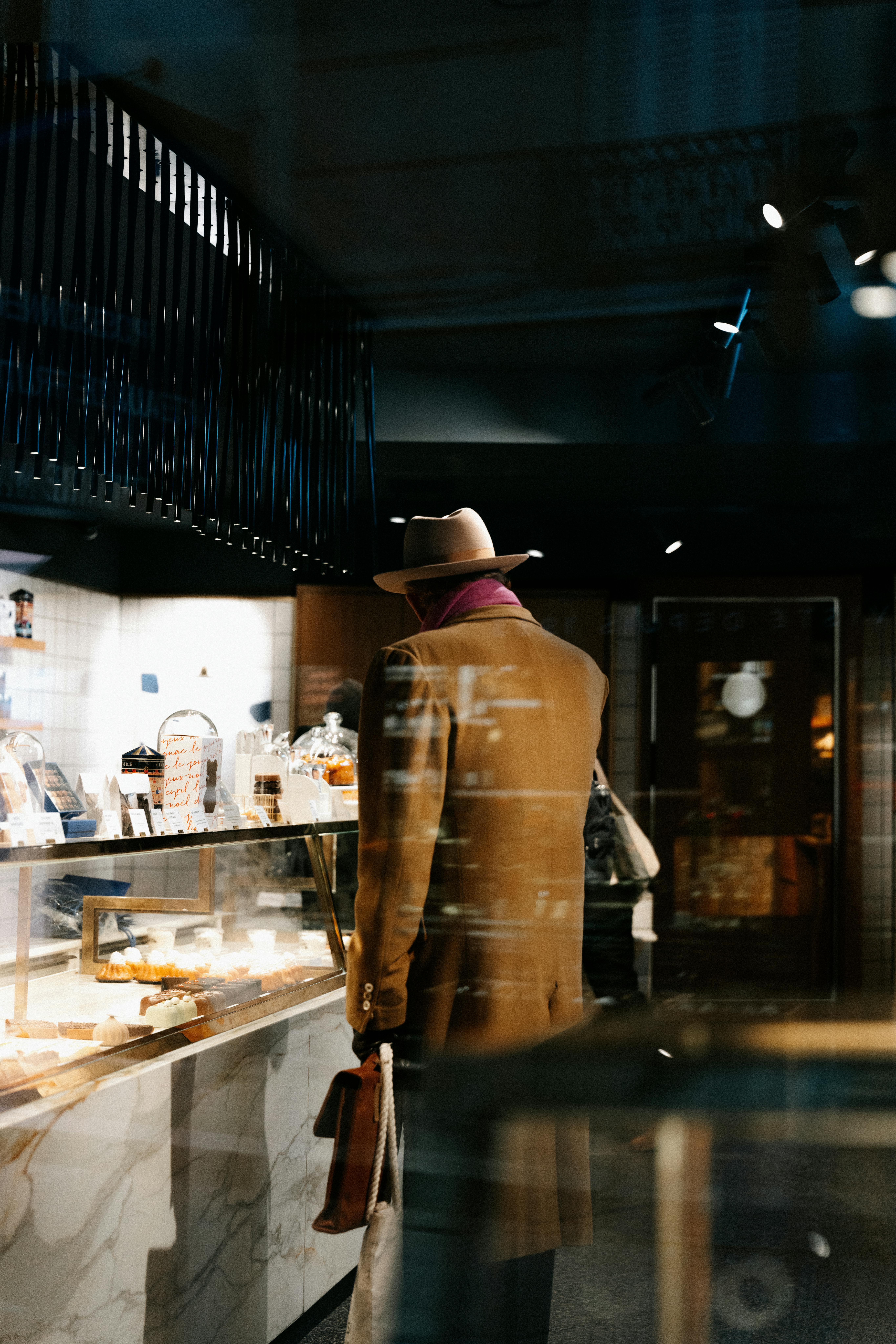 Stylish man with coat and hat at pastry counter through window reflection.