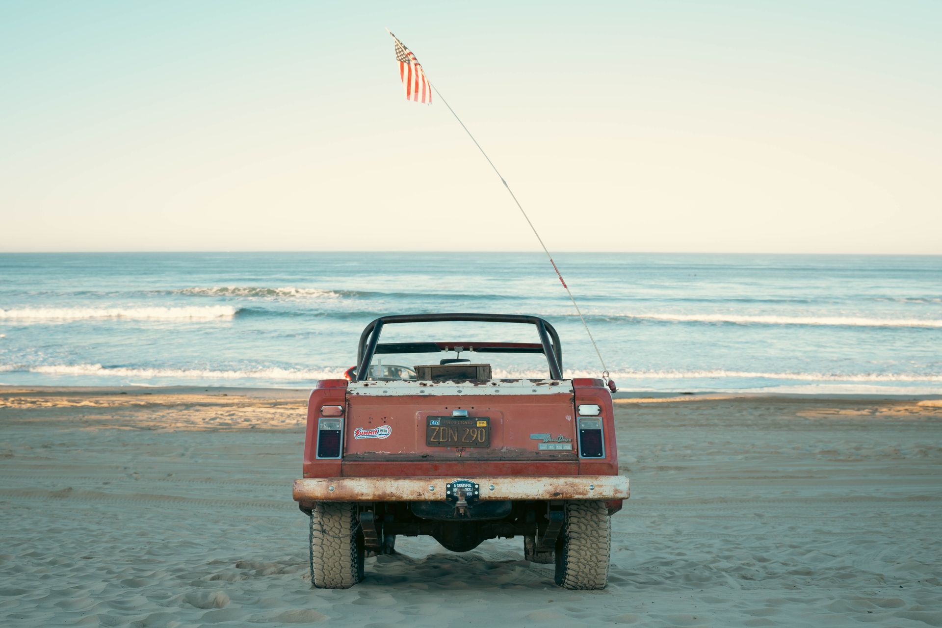A vintage Jeep with an American flag on a sandy California beach during the day.