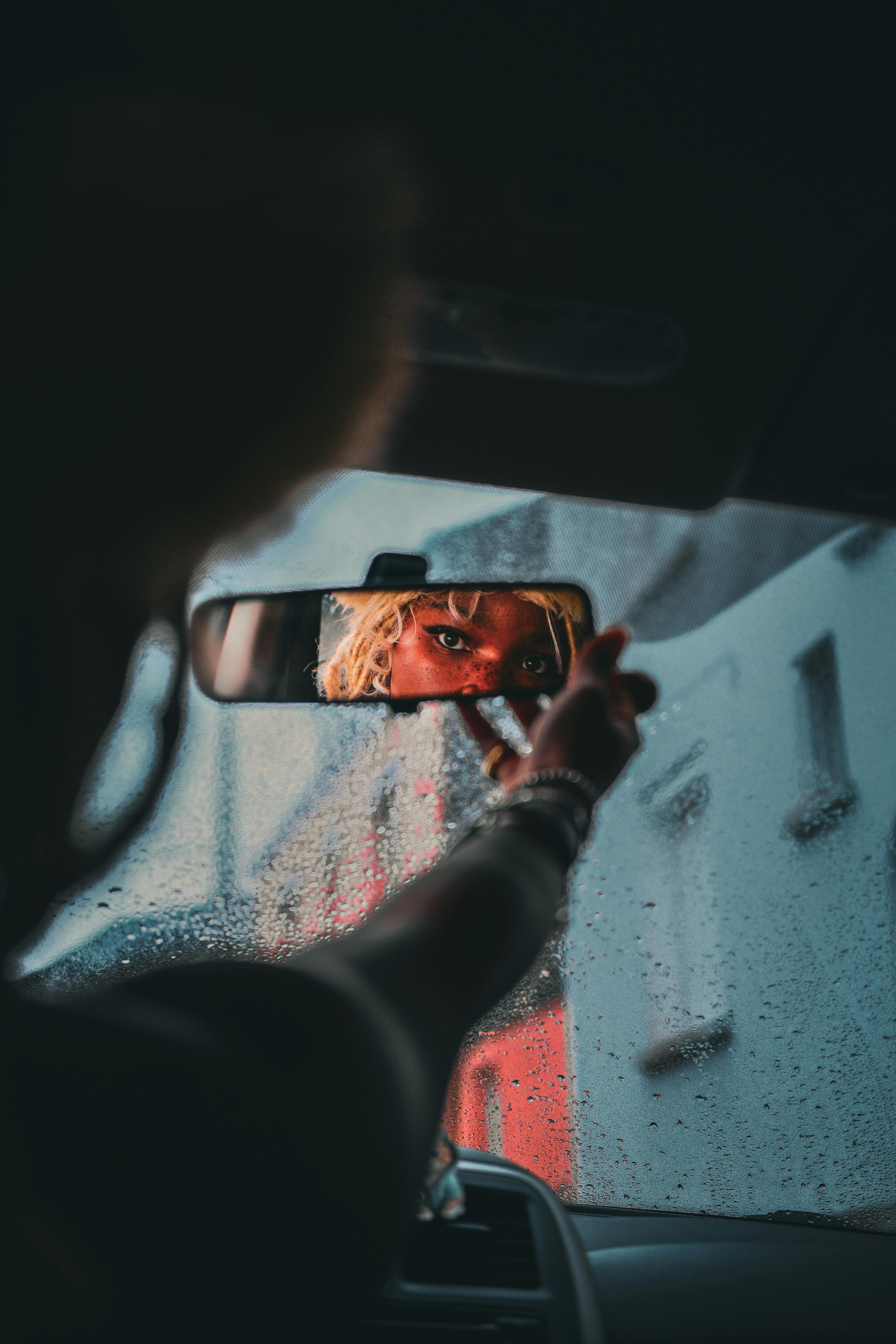 Artistic shot of a person's face in a car mirror with rain on the window.