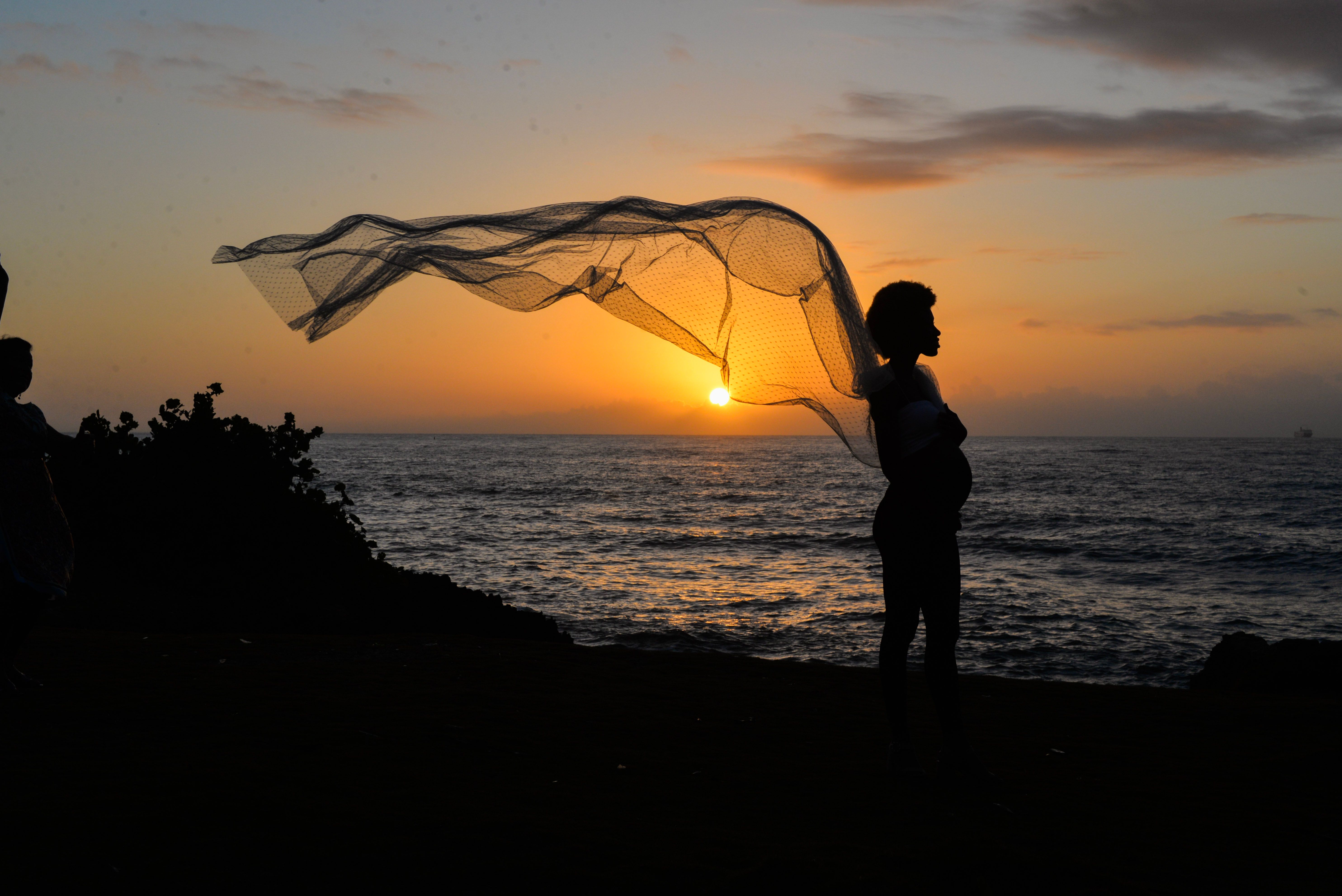Silhouette of a pregnant woman with scarf at sunset by the sea. Peaceful and contemplative scene.