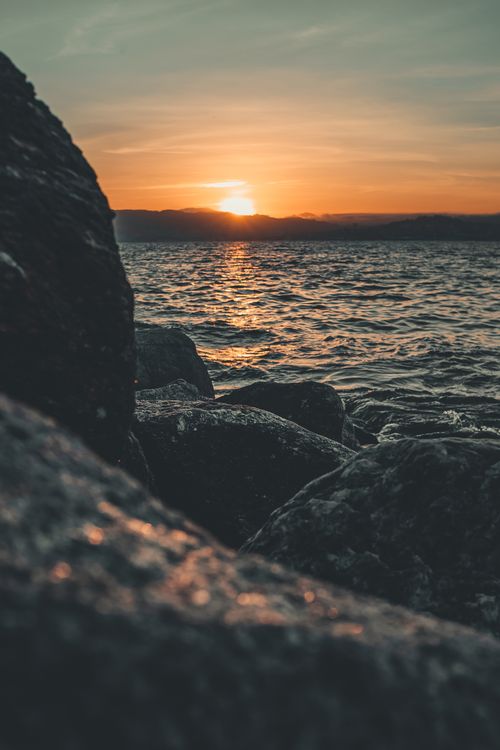 Free Spectacular view of ocean with rippled water near big rough stones with mountains far away under colorful sky at sundown in evening Stock Photo