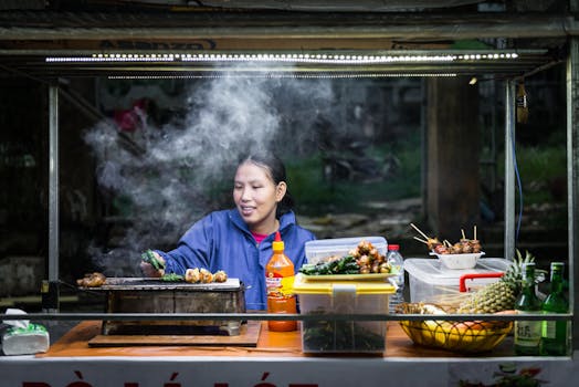 A street food vendor grills skewers amidst steam at a vibrant Hoi An market.