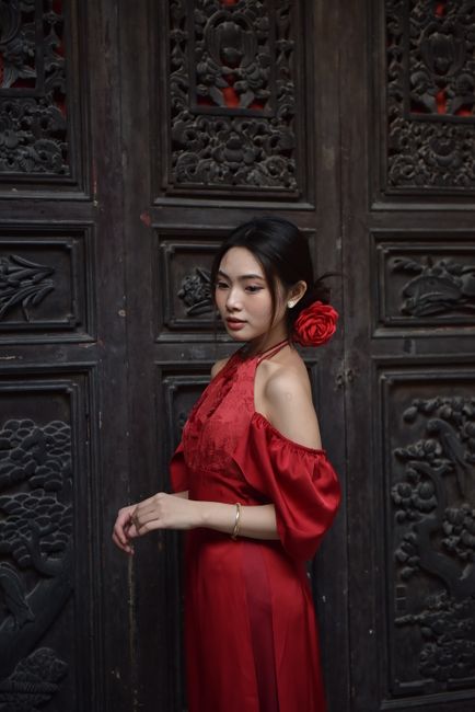 Asian woman in a red dress stands by a decorative wooden door. Elegant pose and intricate design create a classic ambiance.