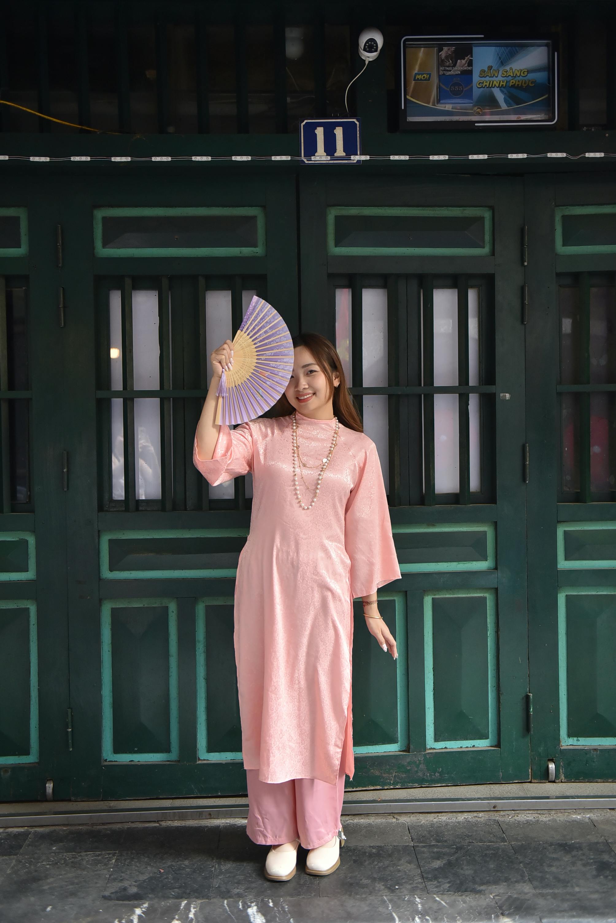A woman in a pink traditional dress poses elegantly with a fan in front of a green door.