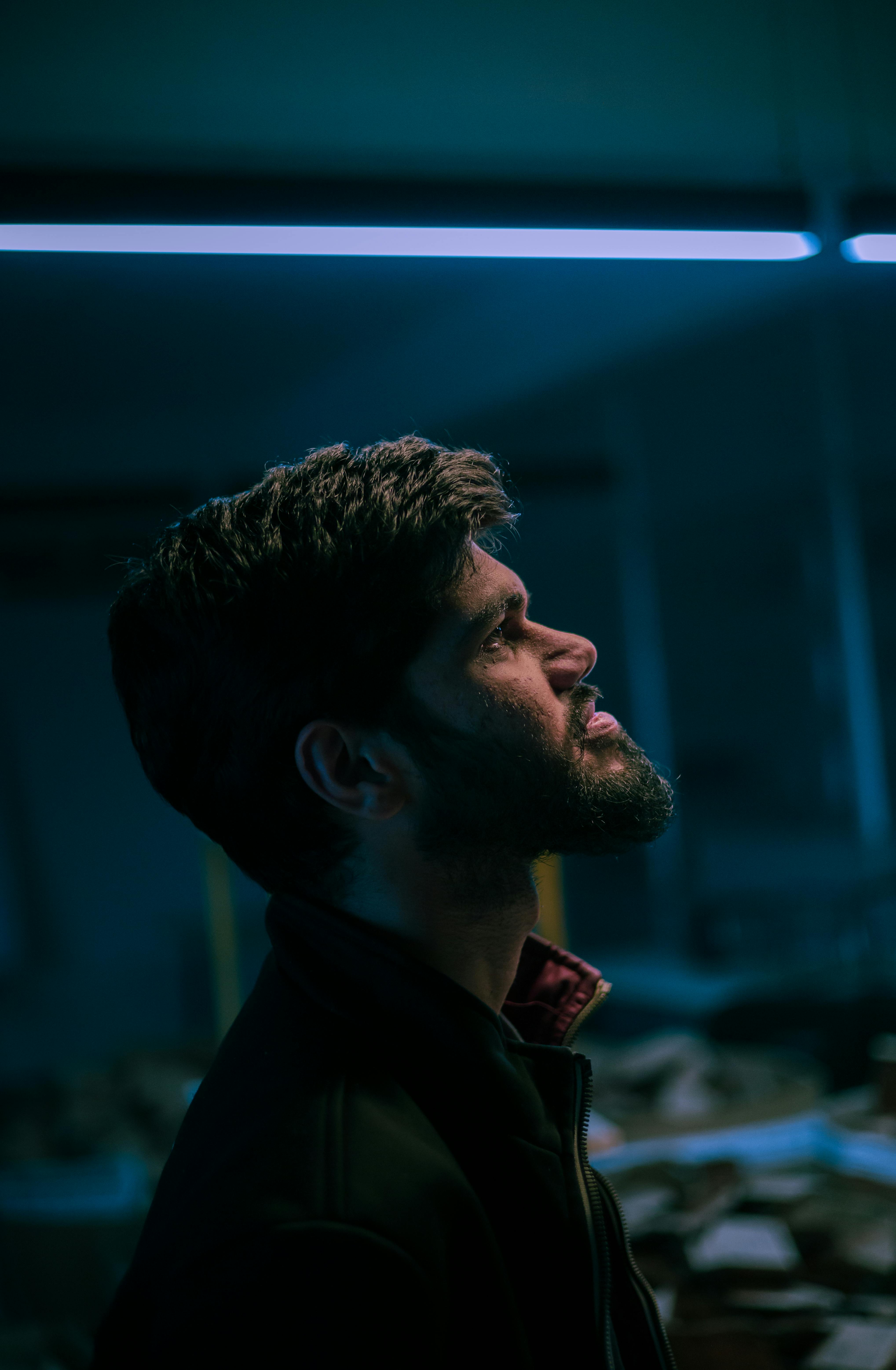 Profile portrait of an adult man in a dark room illuminated by blue neon lighting.