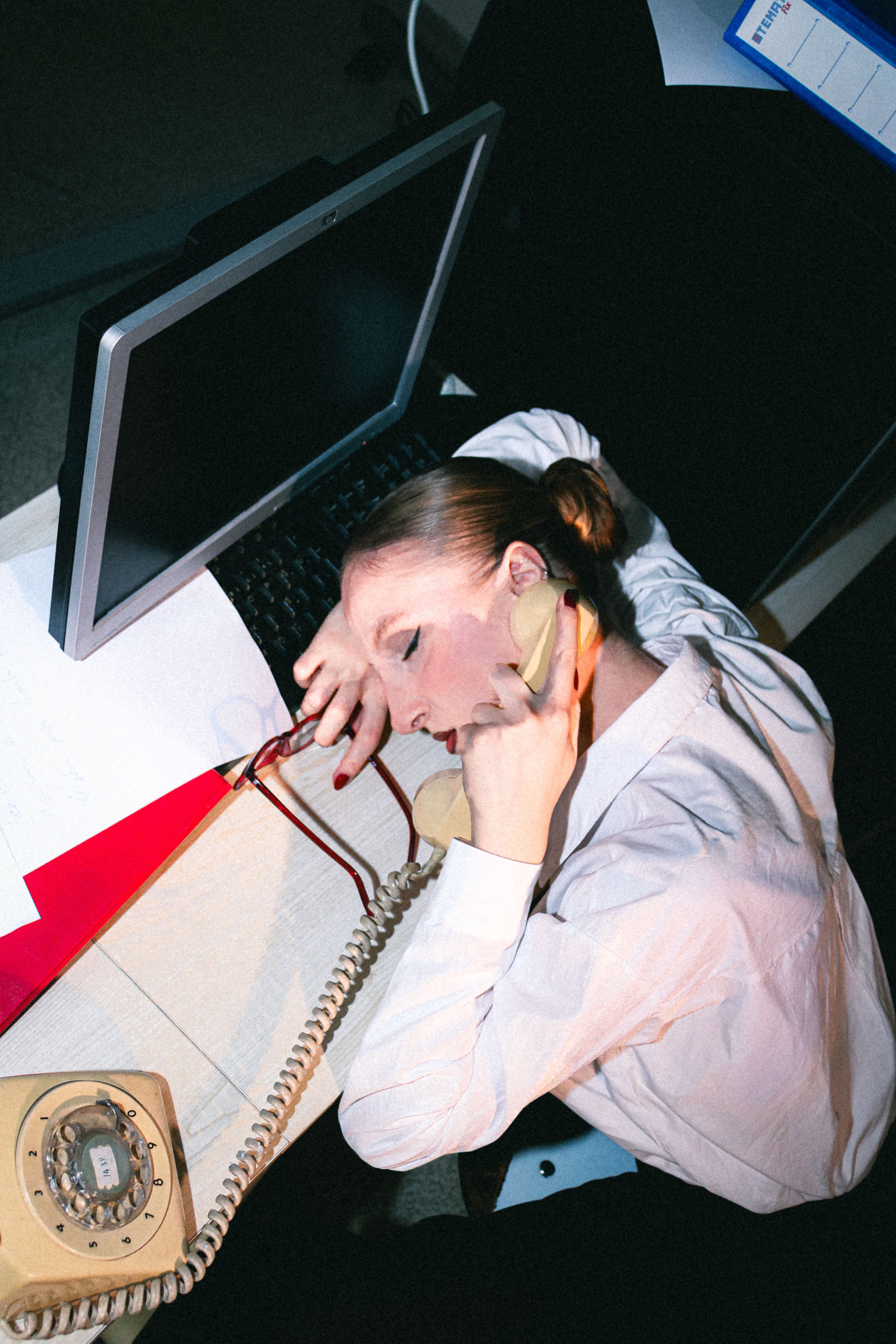 Overworked woman asleep at a desk using a vintage phone, emphasizing workplace stress.