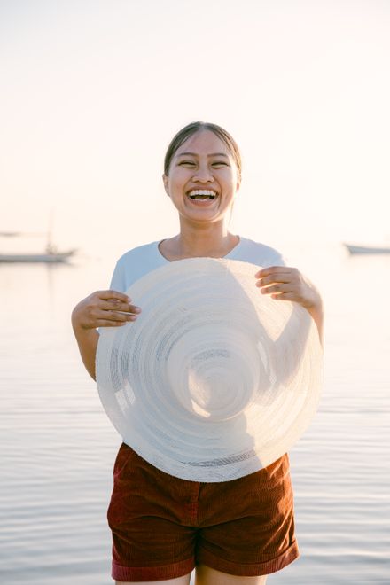 Woman laughing while holding a wide sun hat by the sea — photo by Arjun Adinata on Pexels