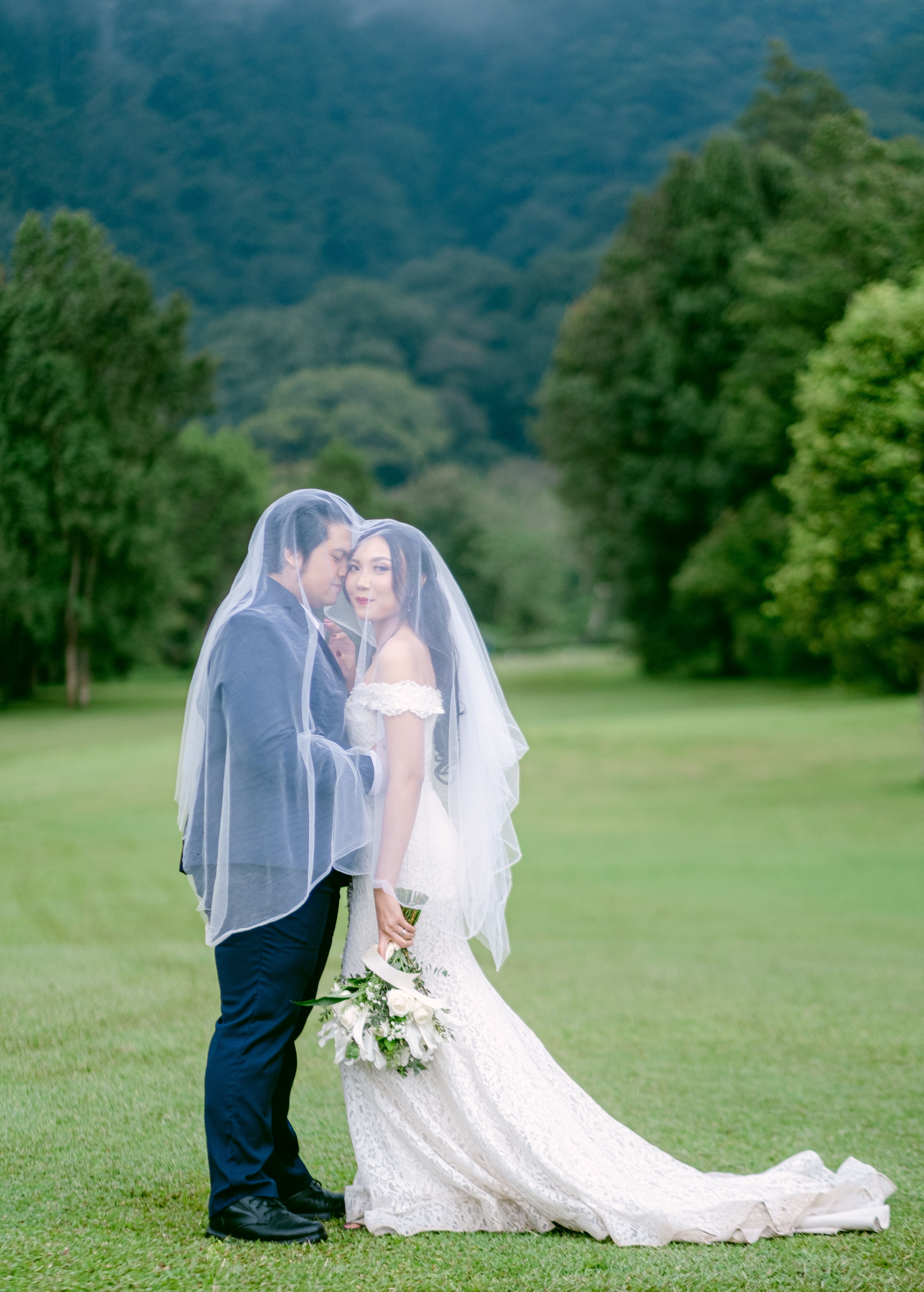 A romantic wedding portrait of a bride and groom under a veil in a green landscape.