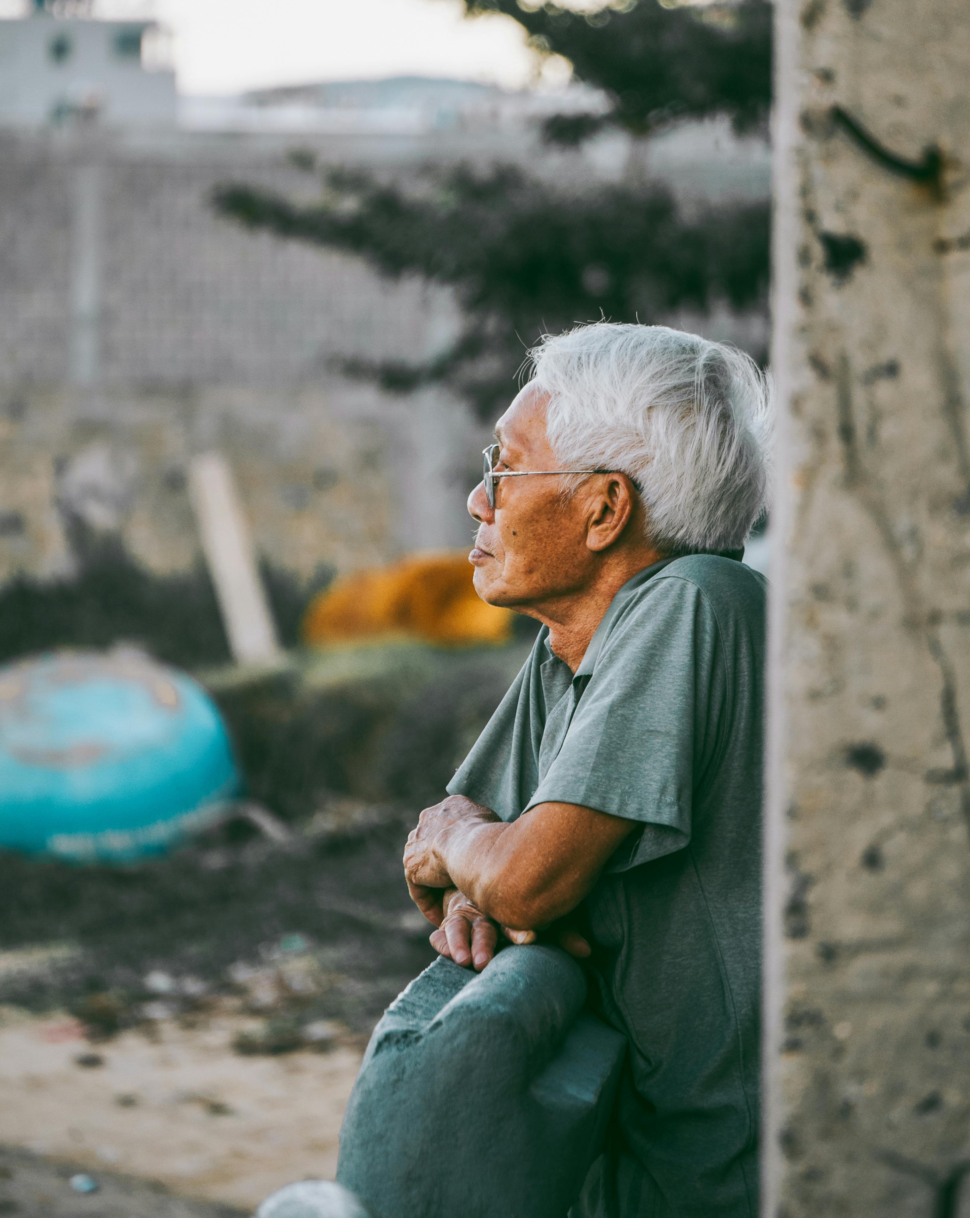 A contemplative elderly man with glasses leans on a wall in Quy Nhơn, Vietnam, displaying a moment of reflection.
