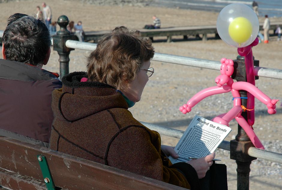Couple sits on beachside bench solving puzzle with balloon animal decoration nearby.