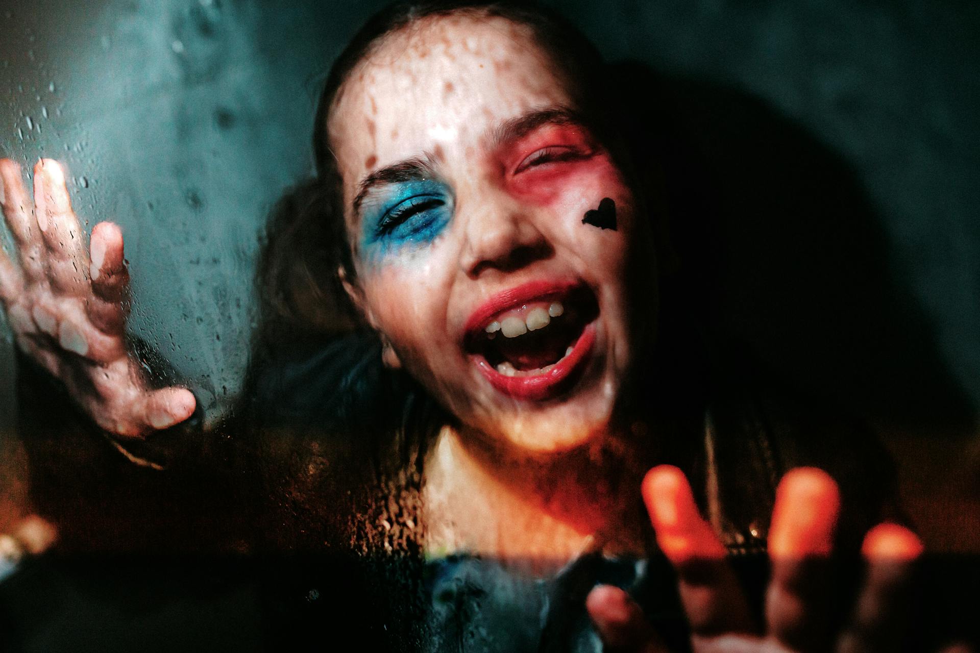 A smiling child with a painted face expressing joy behind a rain-soaked glass indoors.