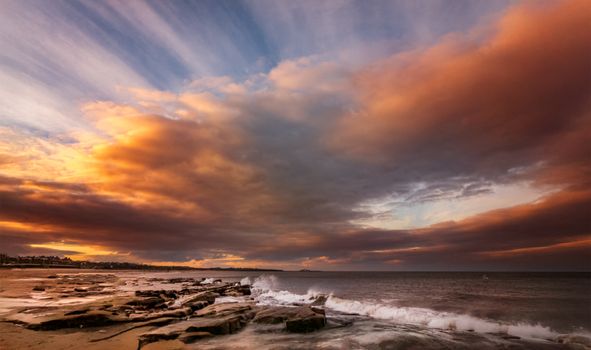 A stunning coastal sunset with vibrant clouds, rocky shoreline, and crashing waves.