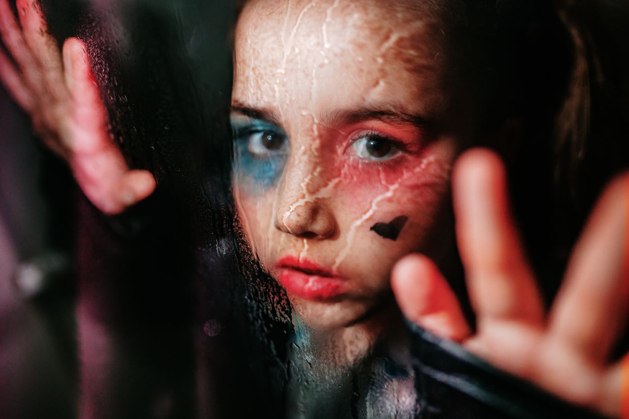 portrait of a child with painted face behind a glass with raindrops ev