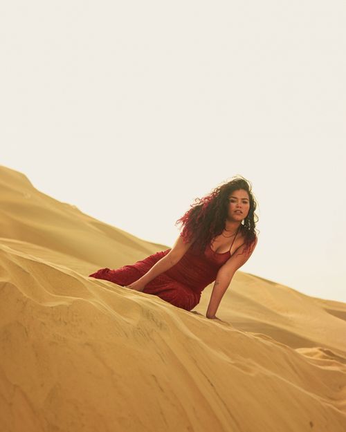 Free A woman in a red dress poses gracefully on rolling golden sand dunes under a bright sky. Stock Photo