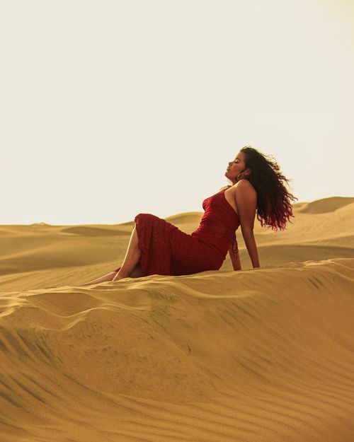 Free A woman in a red dress relaxes on a sunlit sand dune in the desert. Stock Photo