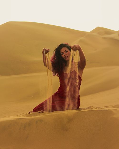 Free A woman in a red dress kneeling in a sandy desert, holding sand in her hands. Stock Photo