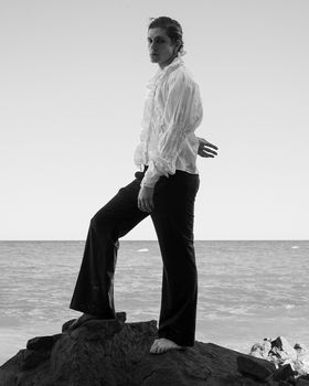A stylish adult stands on rocky shore by the ocean, exuding confidence.
