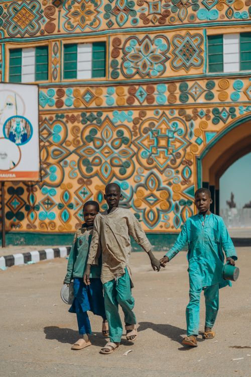 Free Three children in colorful attire walk past a vibrant, patterned building facade on a sunny day. Stock Photo